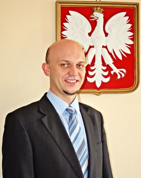 Zdjęcie mężczyzny w niebieskiej koszuli, krawacie w paski i czarnej marynarce, w tle godło Polski